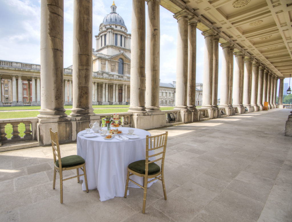A table set for an elegant afternoon tea, complete with a white tablecloth, fine china, and a selection of cakes and sandwiches, is placed under a classical colonnade. The backdrop reveals a historic building with a domed structure, suggesting this might be a prestigious location, possibly a university or a historical institution. The architecture is grand, featuring tall columns and a clear sky above, providing a serene and luxurious ambiance for the tea experience.