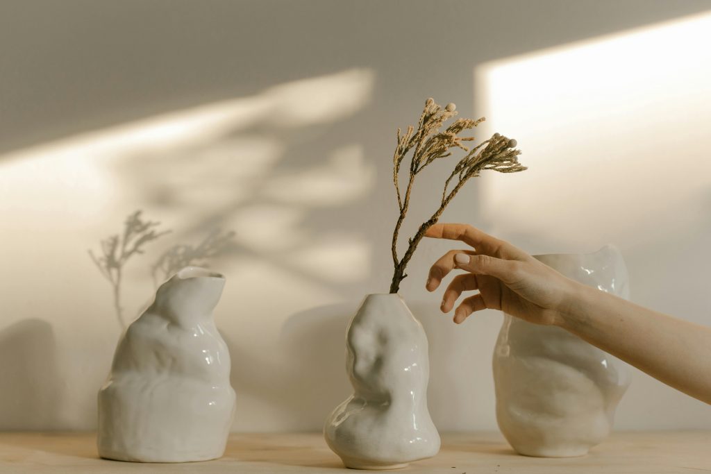 A hand gently placing a dried plant into a white, glossy ceramic vase shaped like a human head and torso. The vase is one of three displayed on a wooden surface, each with unique contours resembling the human form. Soft light casts artistic shadows on the wall behind, enhancing the serene and artistic ambiance of the setting.